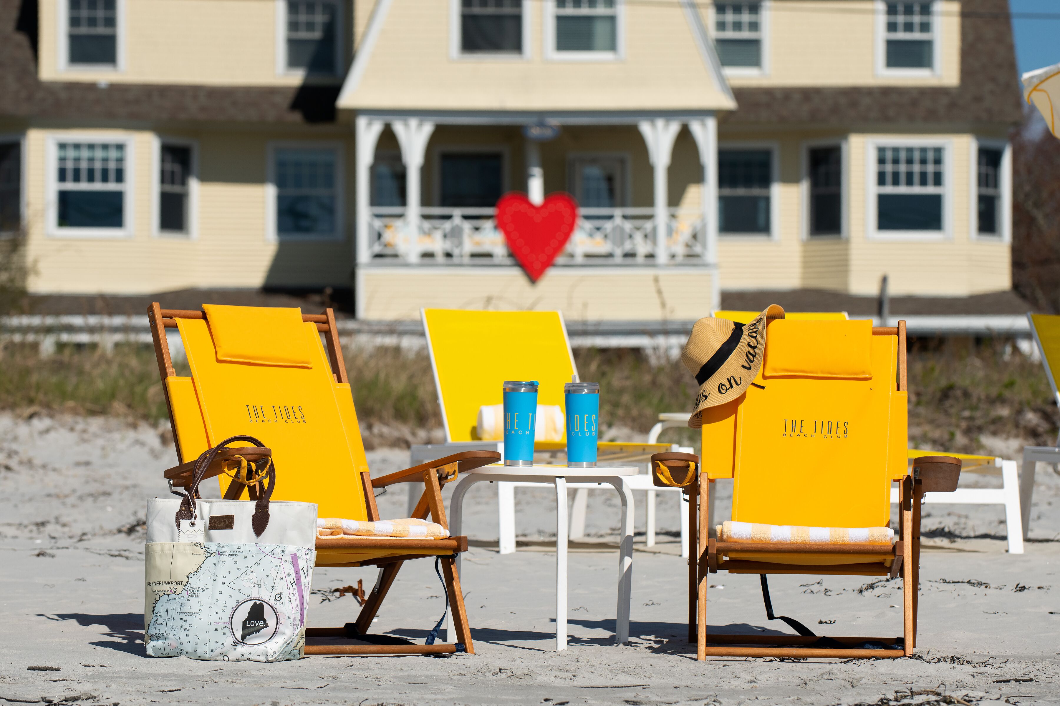 Strandstoelen en een tafel met drankjes en een tas op het zand, met een huis en een rood hart op de achtergrond.