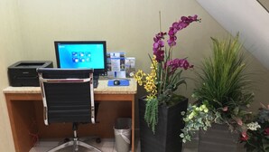 Um espaço de trabalho com um computador de mesa, uma impressora e uma cadeira. Há várias plantas em vasos, incluindo orquídeas e outras folhagens.