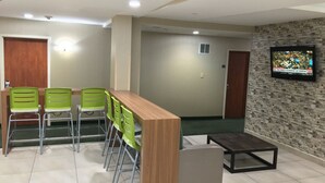 Uma sala de conferências com uma mesa de madeira e cadeiras verdes, uma televisão na parede e uma pequena mesa com um banco.