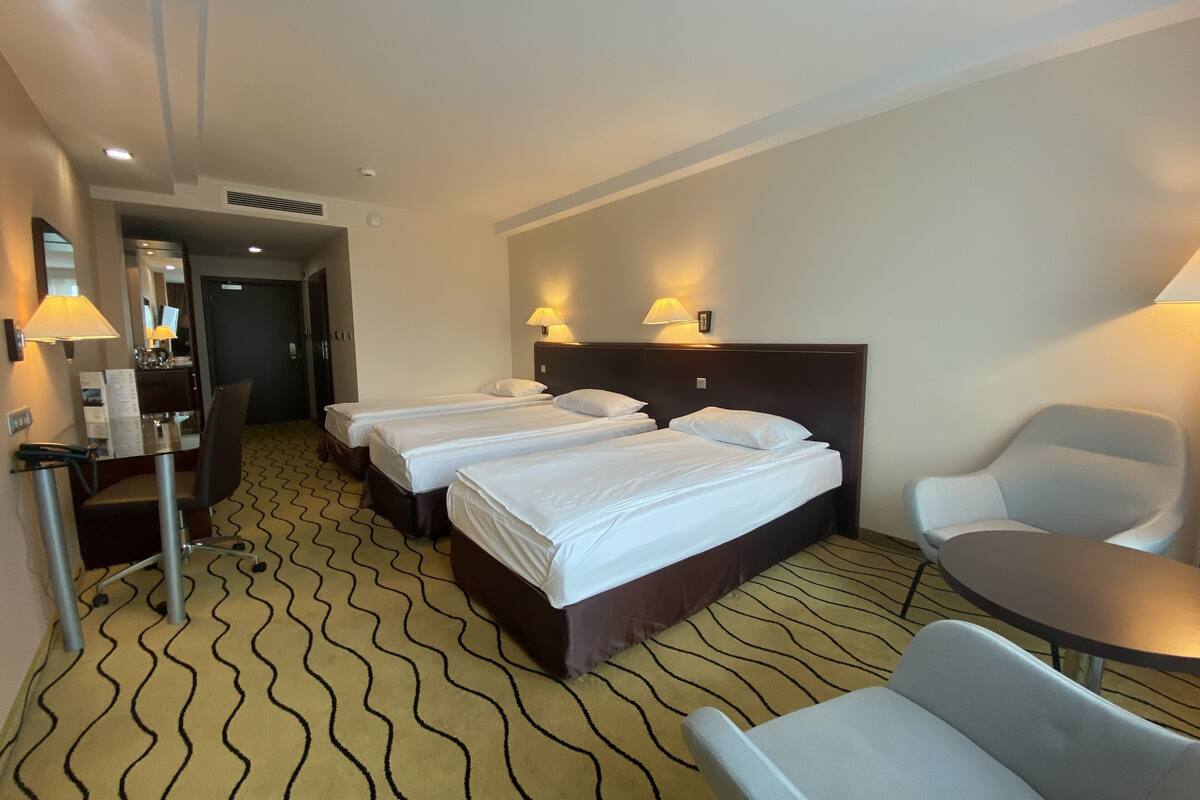 A hotel room with three beds, a desk with a chair, a small table, and two wall-mounted lamps.
