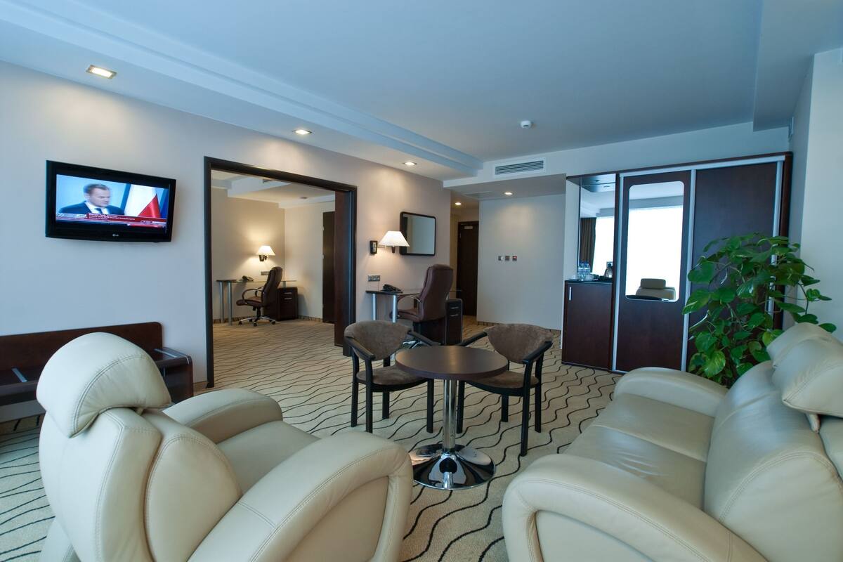 A hotel room with a sofa, a TV mounted on the wall, a table with chairs, a lamp, and a potted plant.