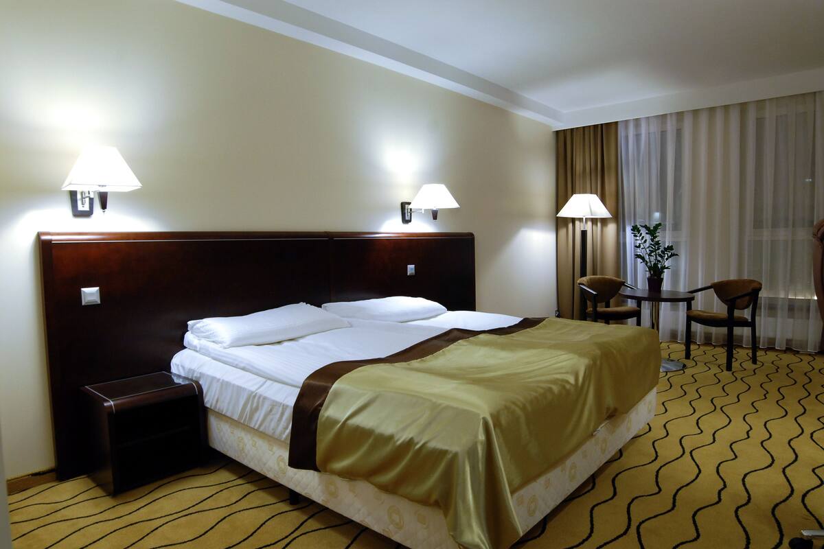 A hotel room with a large bed, two bedside lamps, a small table with chairs, and a window with curtains.