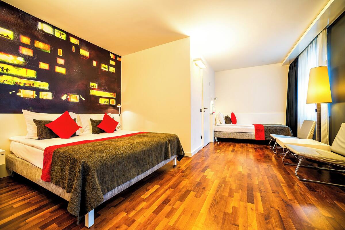 Una habitación de hotel moderna con una cama grande, una cama más pequeña, una zona de estar y un cuadro con una vista urbana.