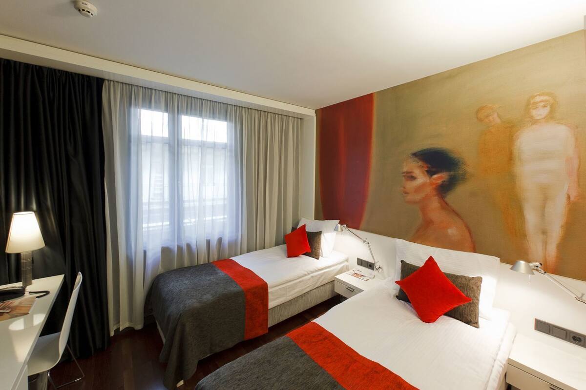 Habitación de hotel con dos camas, un escritorio, una silla, una lámpara y un cuadro en la pared.