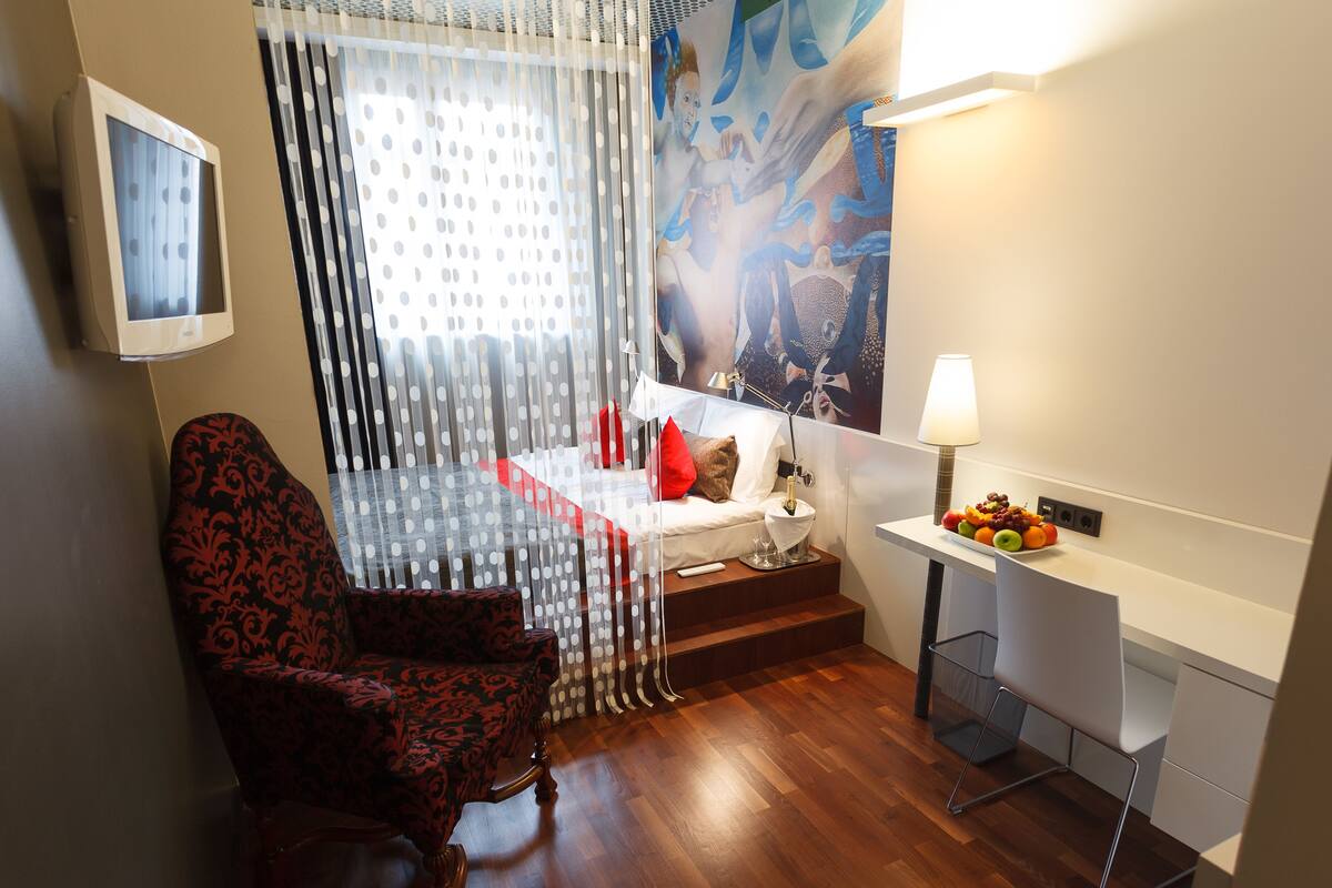 Habitación de hotel con cama, escritorio con lámpara, silla, frutero y un cuadro en la pared.