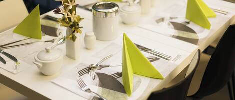 Une table dressée avec de la vaisselle blanche, comprenant des assiettes, des couverts et une théière, agrémentée de porte-serviettes verts et d’un vase avec des fleurs.