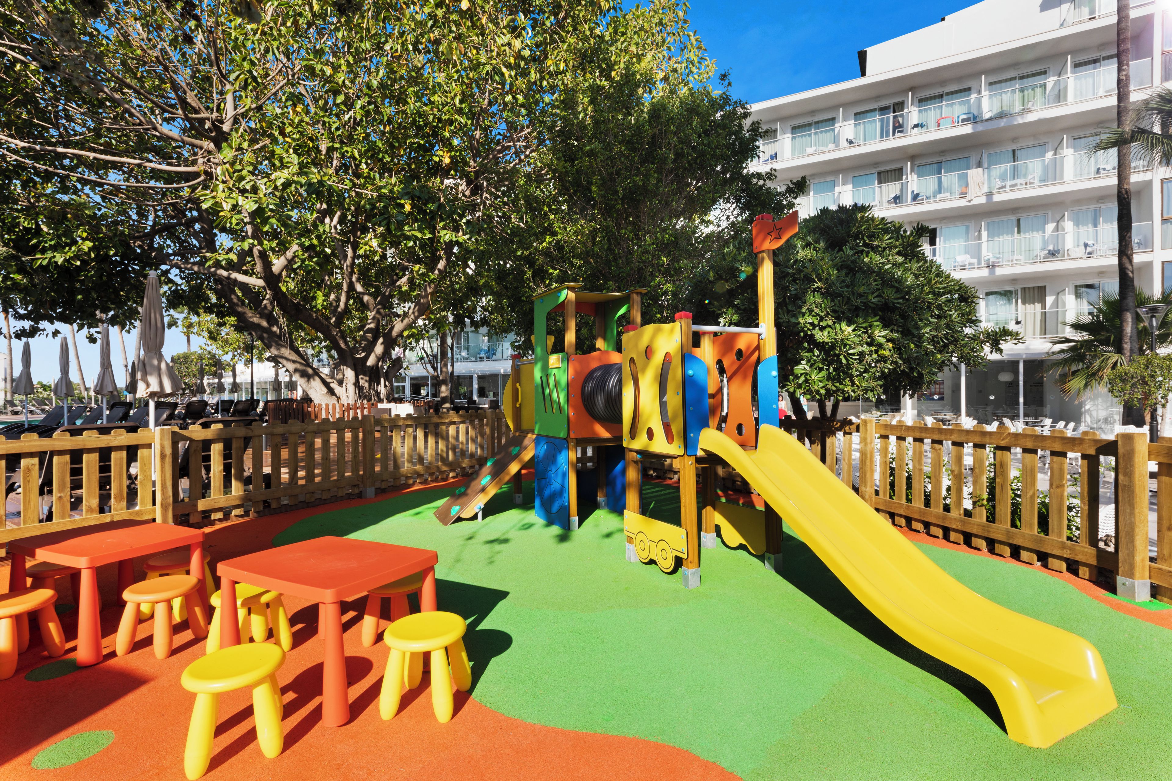 Un parc infantil amb tobogan i taules, envoltat d'una tanca de fusta.