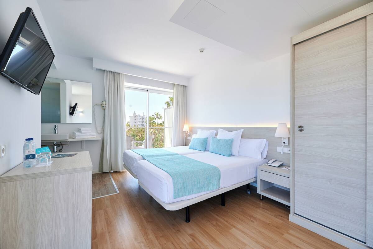 A modern hotel room with a large bed, a TV mounted on the wall, a desk with a phone, and a view of palm trees through the window.