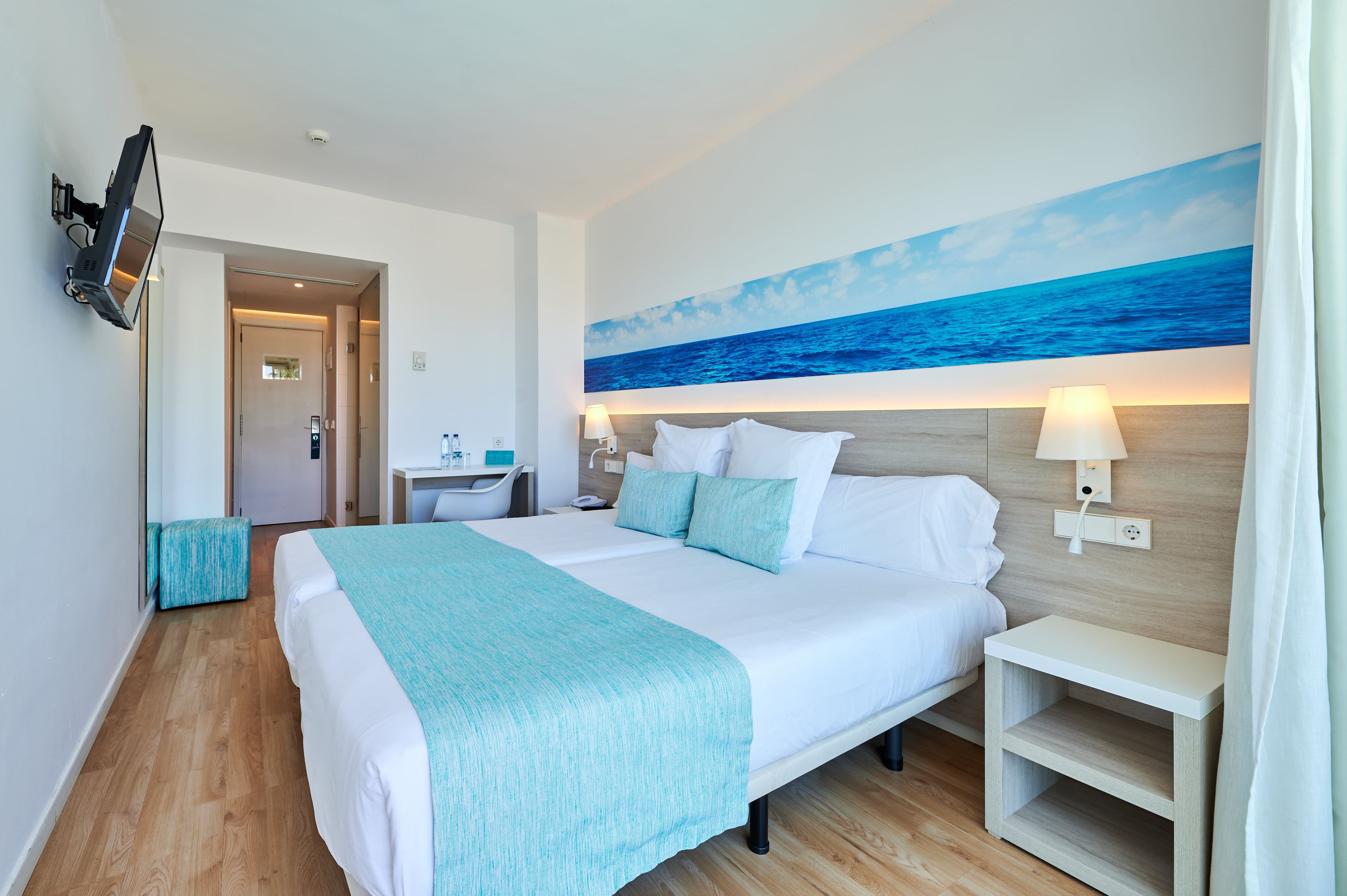 A hotel room with a large bed, a desk with a chair, a television mounted on the wall, and a painting of the ocean.