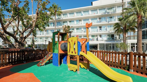 A playground with a yellow slide and a play structure, surrounded by a wooden fence and palm trees, with a hotel building in the background. - Alua Boccaccio (Alcúdia)