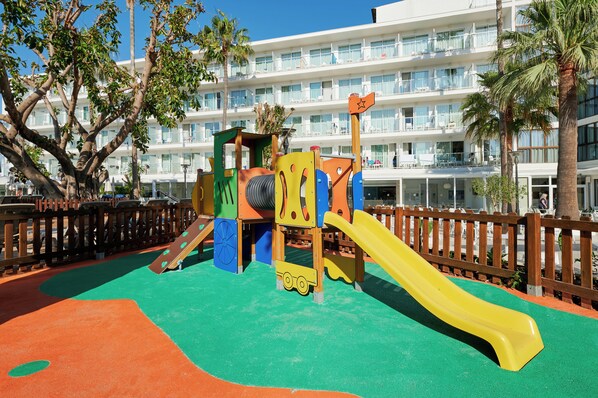 A playground with a yellow slide and a play structure, surrounded by a wooden fence and palm trees, with a hotel building in the background. - Alua Boccaccio (Alcúdia)