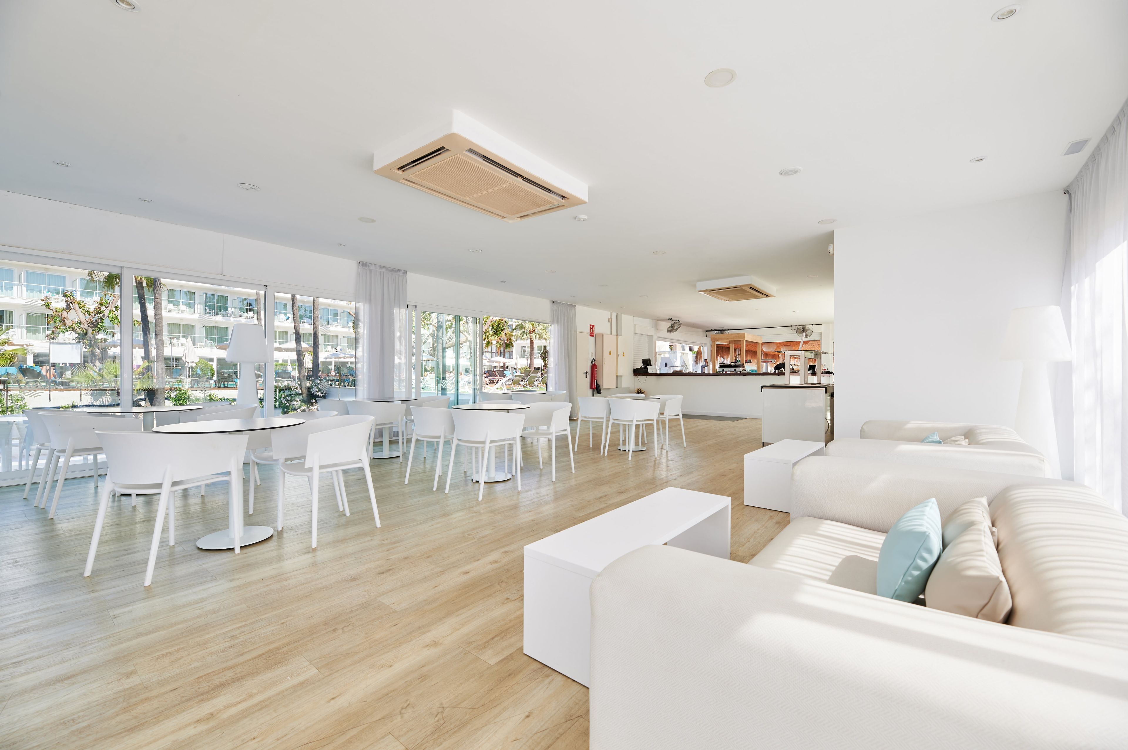 A spacious, modern living area with a large dining table, white furniture, and large windows offering a view of the outside.