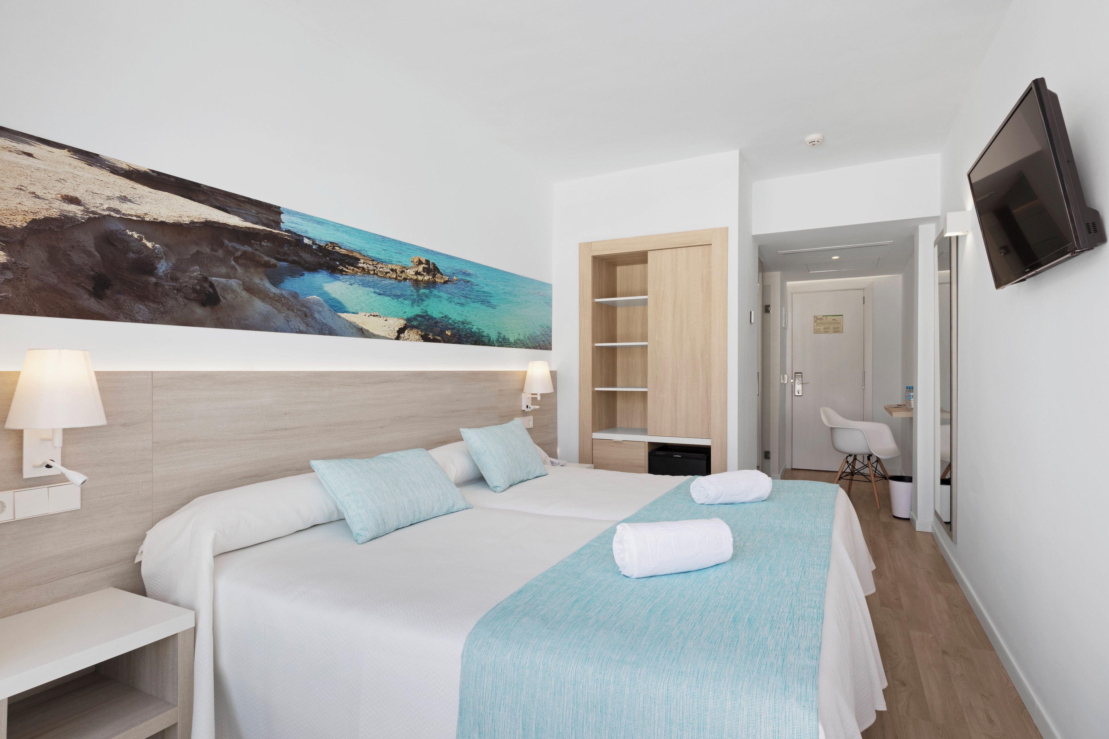 Habitación de hotel con cama, escritorio, silla, televisor y un cuadro de una playa.