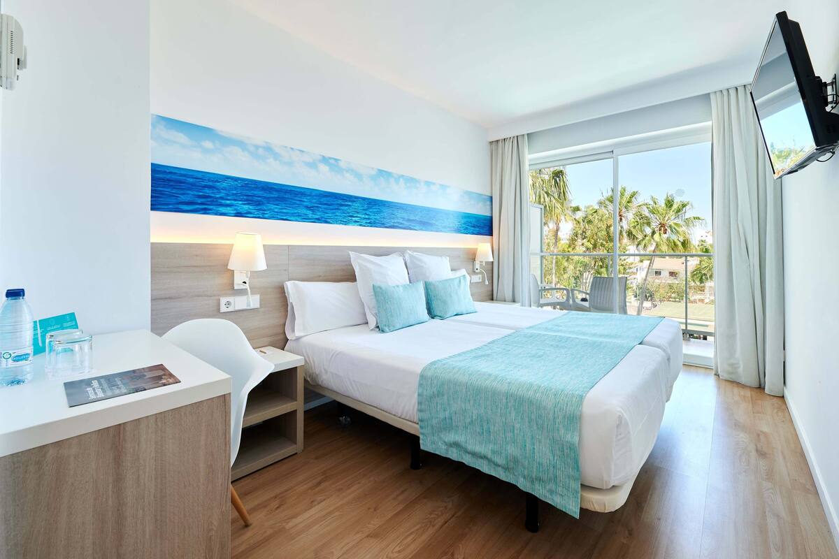 A hotel room with a bed, a desk, a chair, a TV, and a large window with a view of palm trees.