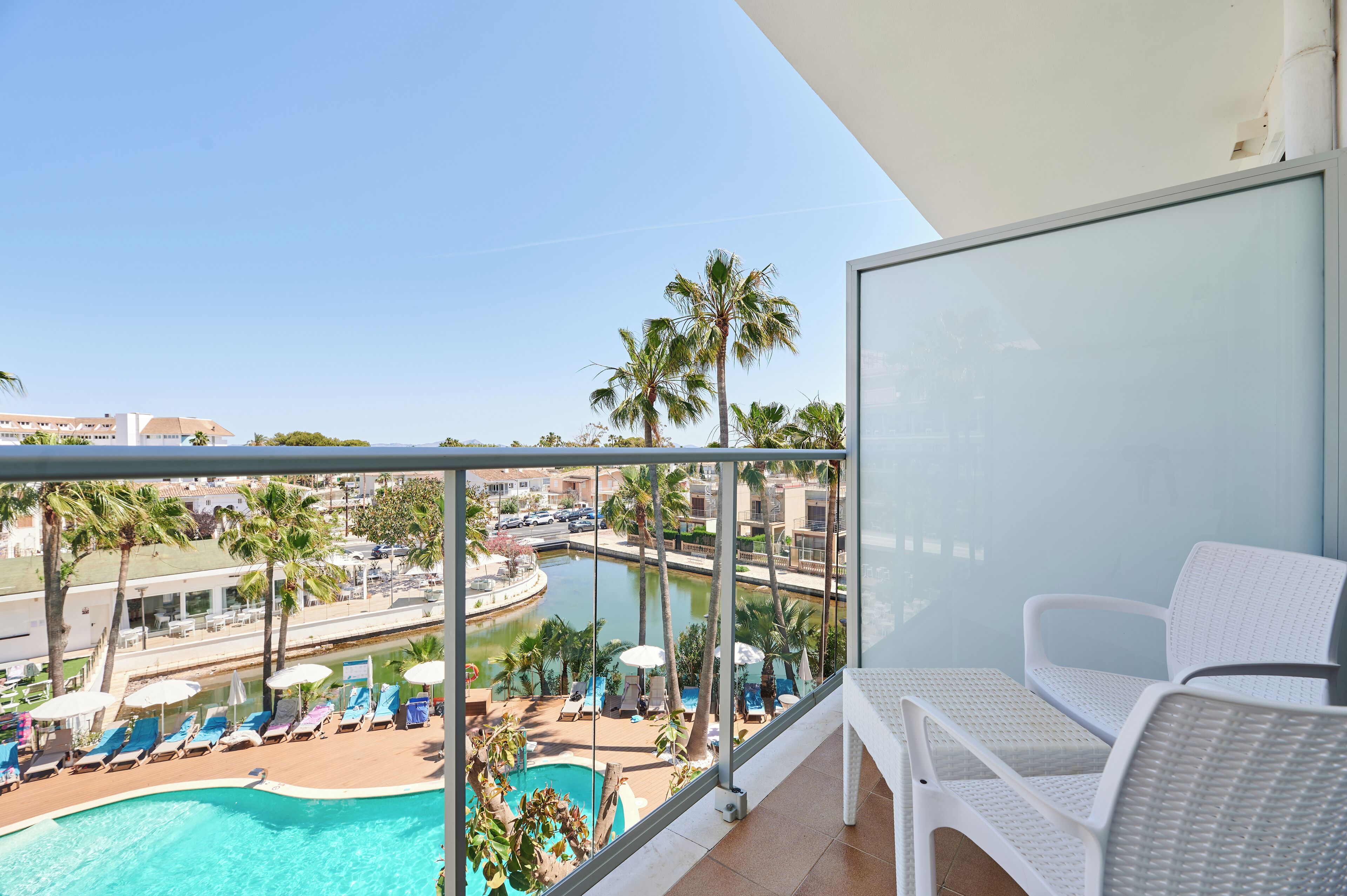Un balcone con vista sulla piscina, sedie in vimini bianco e cielo sereno.