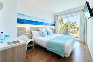 Habitación de hotel con cama, escritorio, silla, televisor y un amplio ventanal con vista a palmeras.