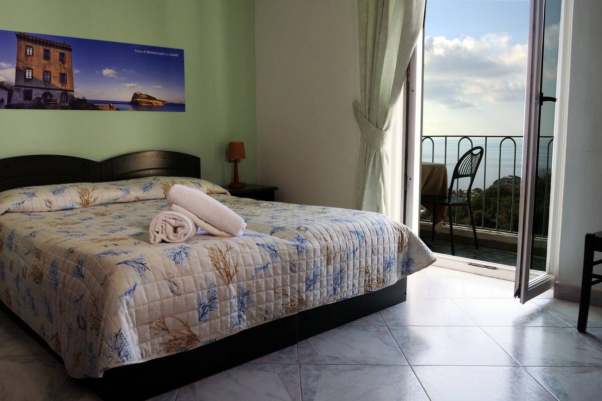 Ein Schlafzimmer mit einem Bett, zwei Nachttischen, einem Fenster mit Meerblick und einem Bild an der Wand.
