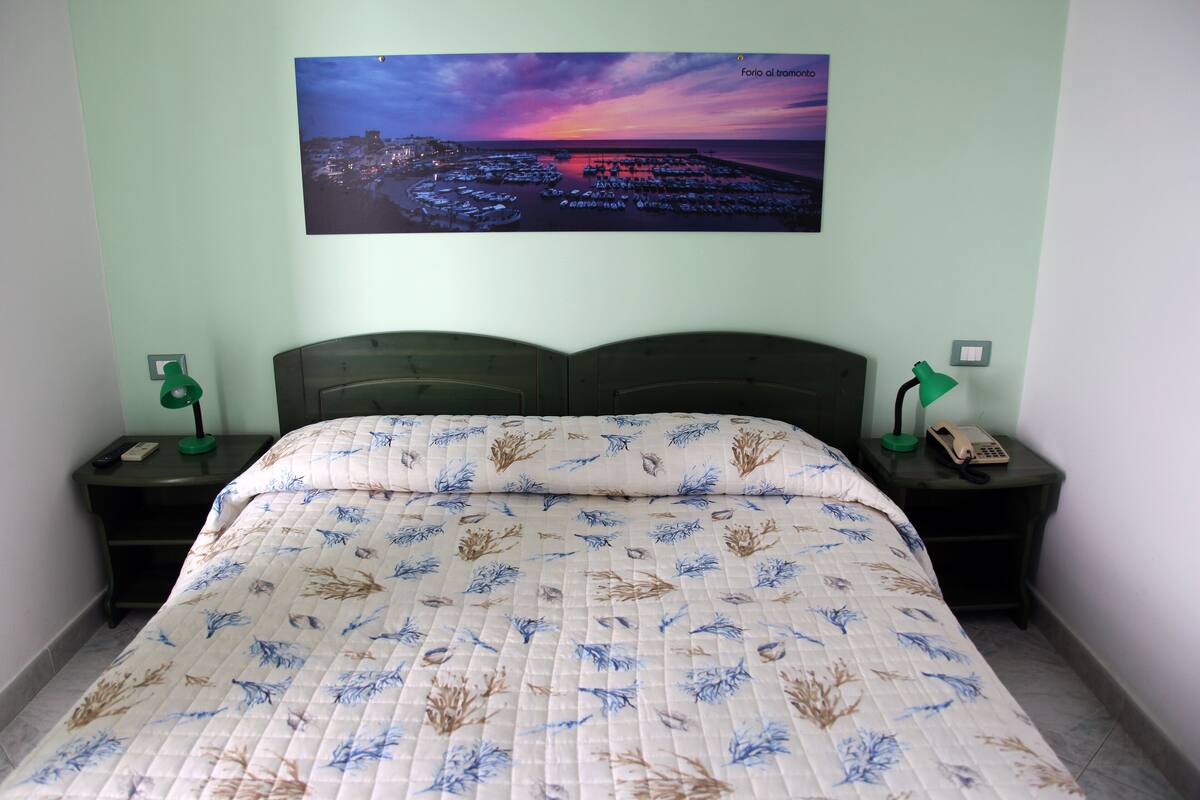 Ein ordentlich bezogenes Bett mit floralem Überwurf, zwei Nachttische mit Lampen und Telefonen sowie ein Stadtblick an der Wand.