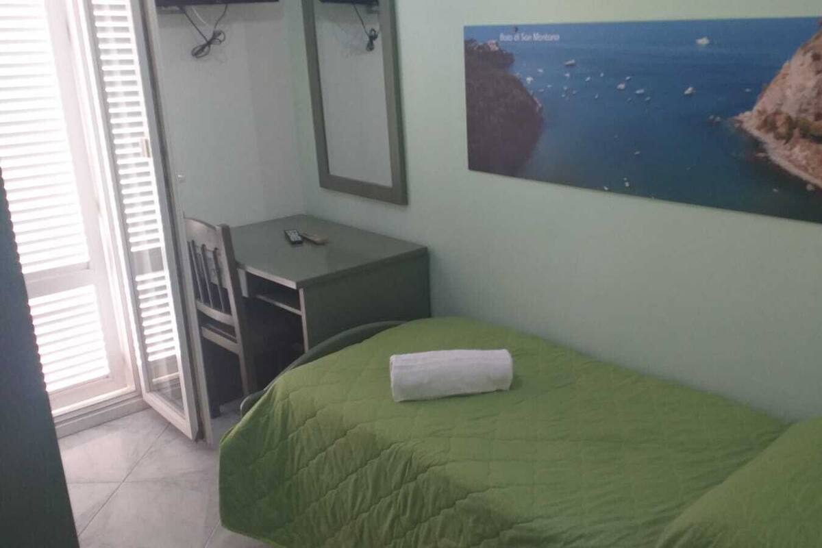 Ein Schlafzimmer mit einer grünen Bettdecke, einem Schreibtisch mit Stuhl, einem Spiegel und einem Wandbild mit einer Küstenlandschaft.