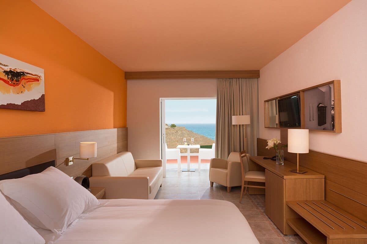 Habitació d'hotel amb un llit gran, dos seients còmodes, una taula de treball amb una làmpada i una televisió. Hi ha una finestra amb vista al mar.