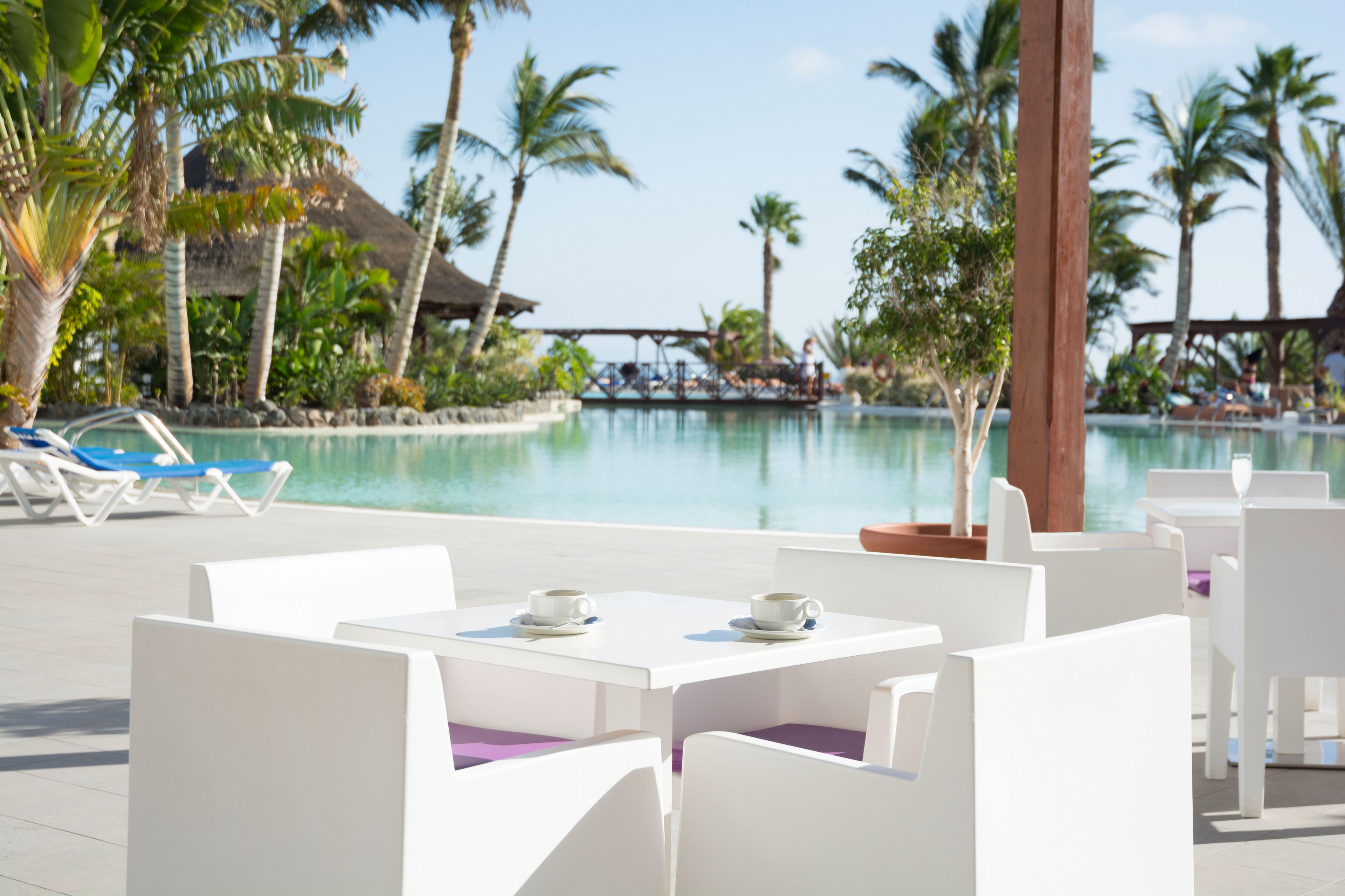 A poolside area with white lounge chairs and tables, palm trees, and a thatched-roof structure in the background.