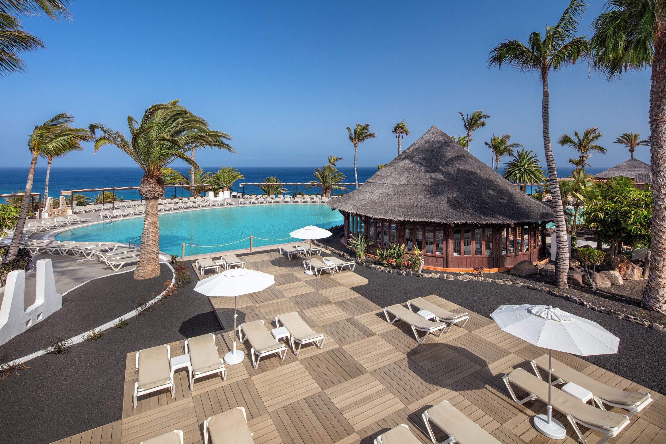 A resort pool area with lounge chairs, umbrellas, and a thatched-roof building.