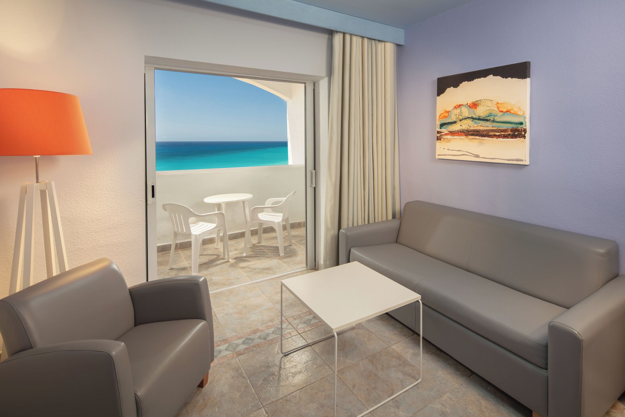 A hotel room with a grey sofa, a small white table, and a view of the ocean through a glass door.