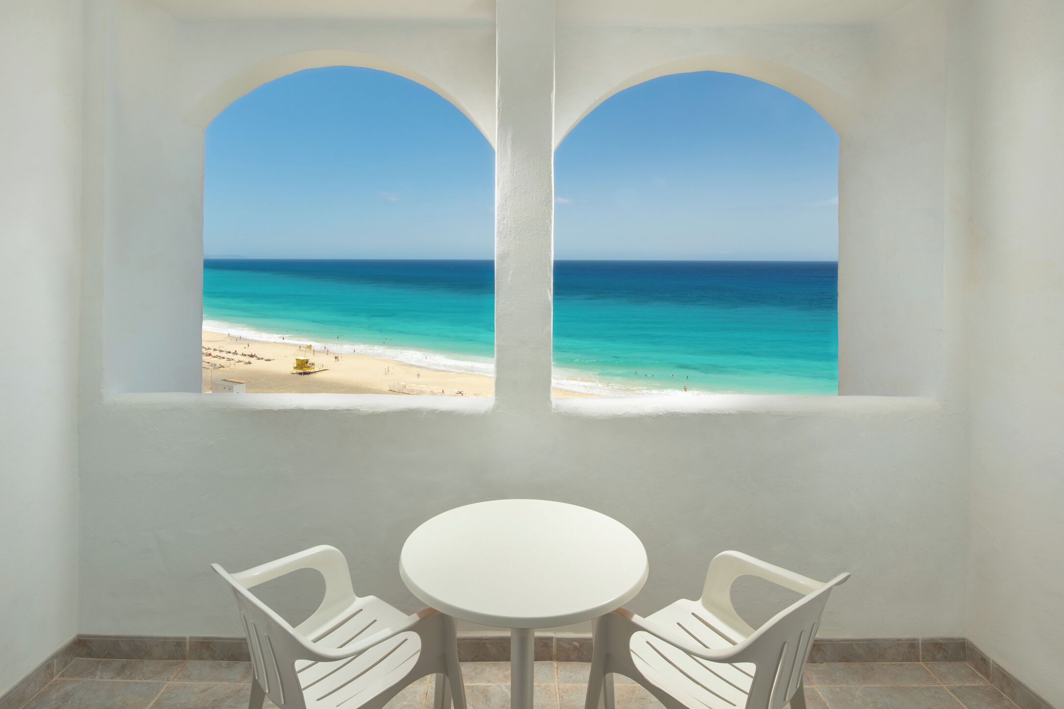 Een kamer met uitzicht op een strand door twee ronde ramen, een ronde tafel en twee witte stoelen.