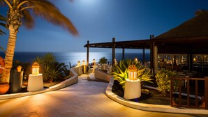 A nighttime outdoor area with a view of the ocean, palm trees, and a covered seating area with tables and chairs. - Club Jandía Princess - All Inclusive (Pajara)