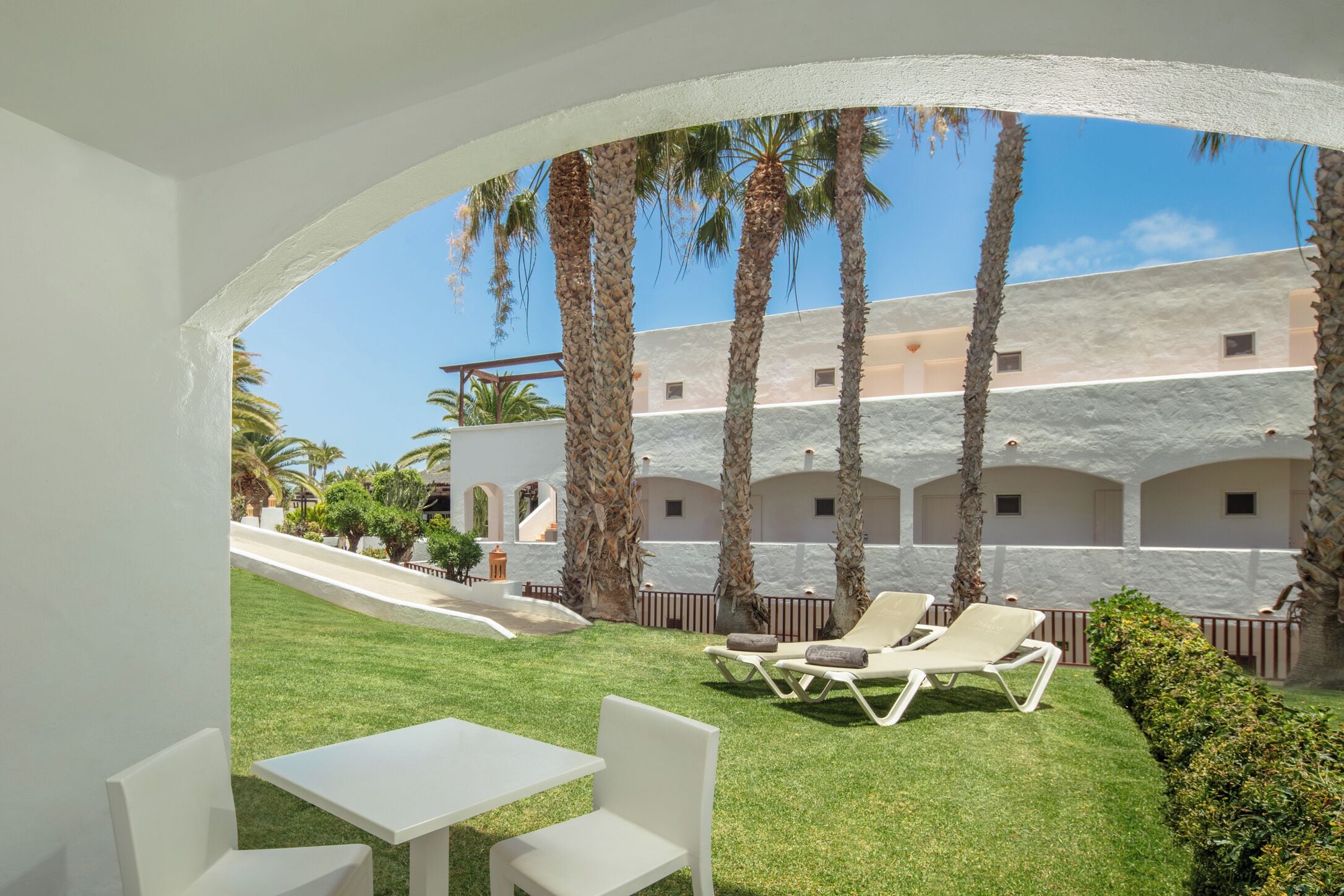 Un patio con una mesa y sillas blancas, palmeras y un edificio con arcos al fondo.