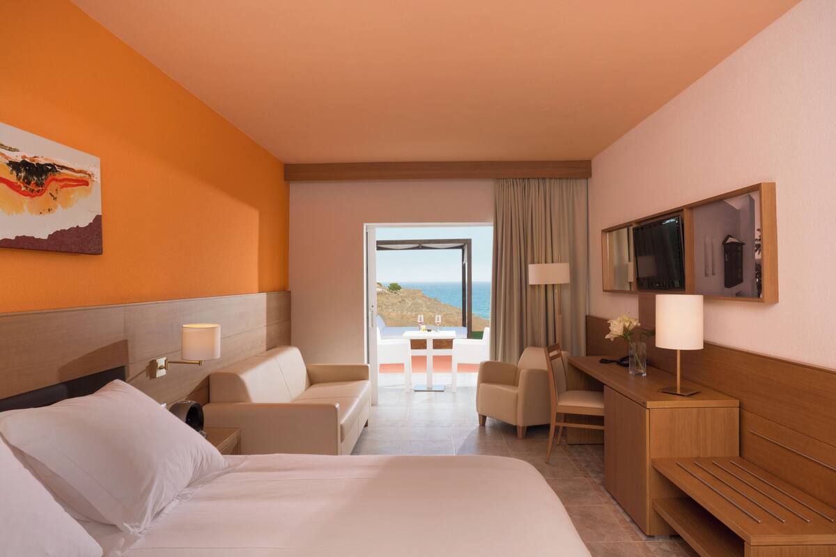 Habitació d'hotel amb llit, dues butaques, escriptori, televisió i vista al mar.
