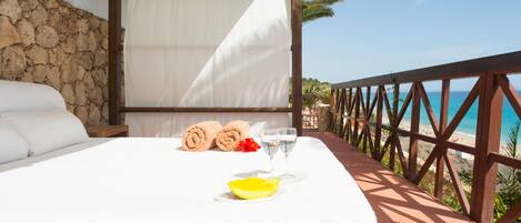 A bed with a white sheet, a glass of water, and a lemon wedge on a wooden platform overlooking a beach.