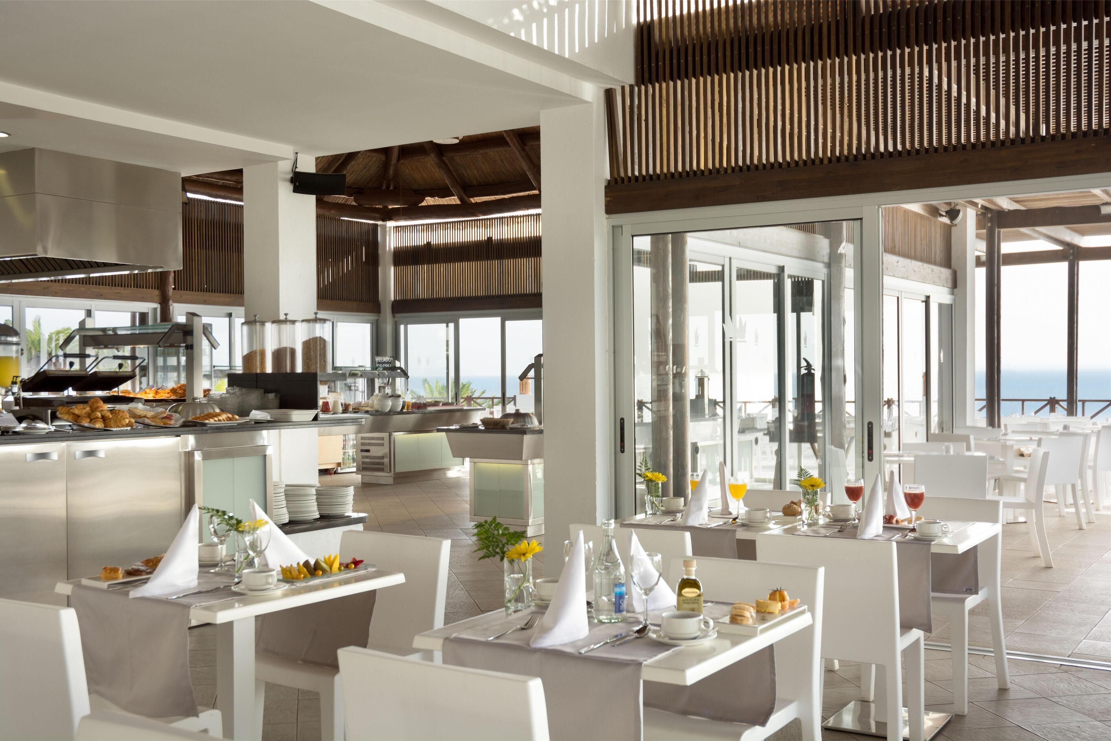 A modern restaurant with white furniture, a buffet area, and large windows offering ocean views.