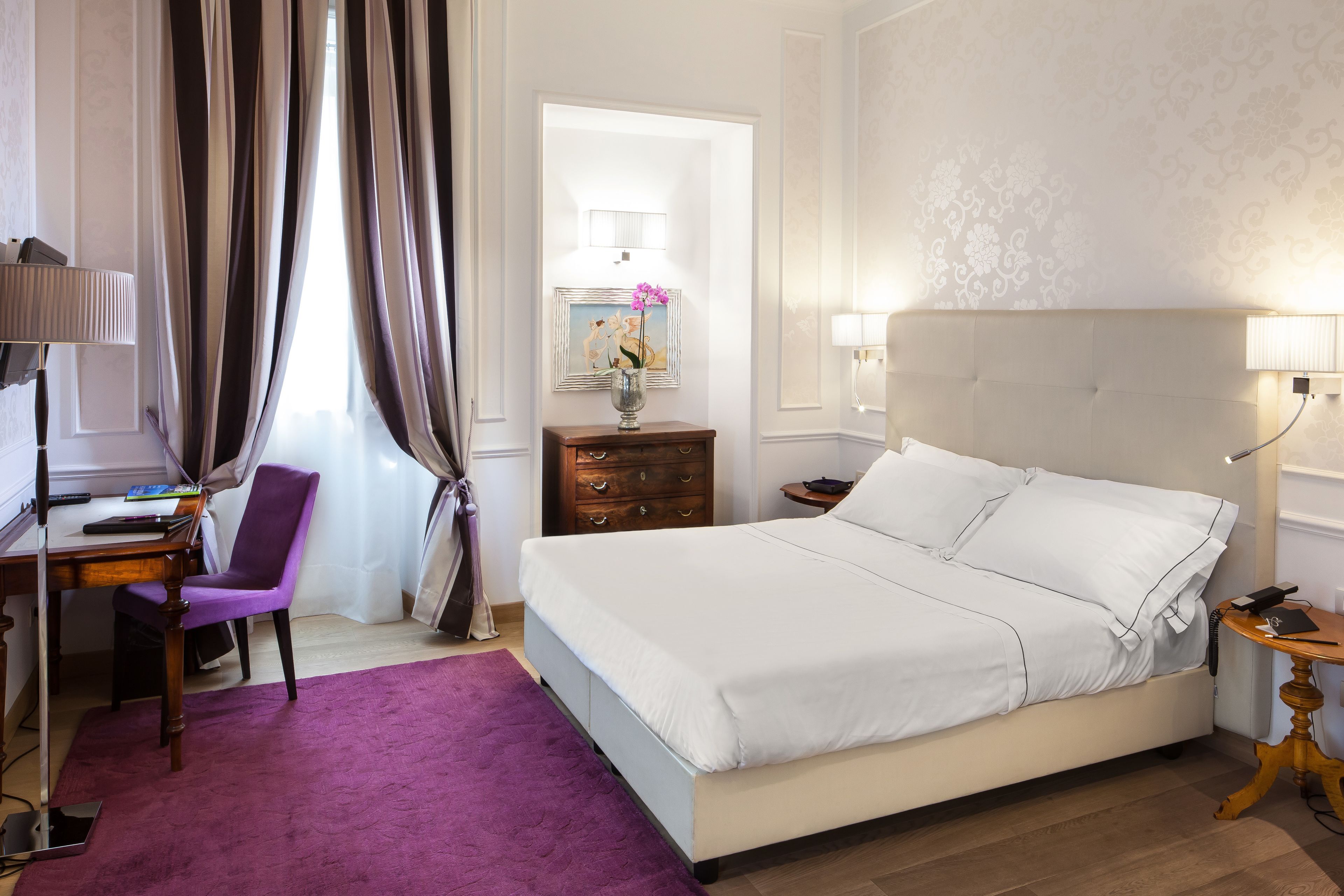 Una habitación de hotel con una cama, un escritorio con silla, un tapete morado, una mesita de noche y una lámpara.