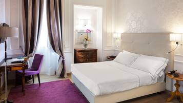 Una habitación de hotel con una cama, un escritorio con silla, un tapete morado, una mesita de noche y una lámpara.