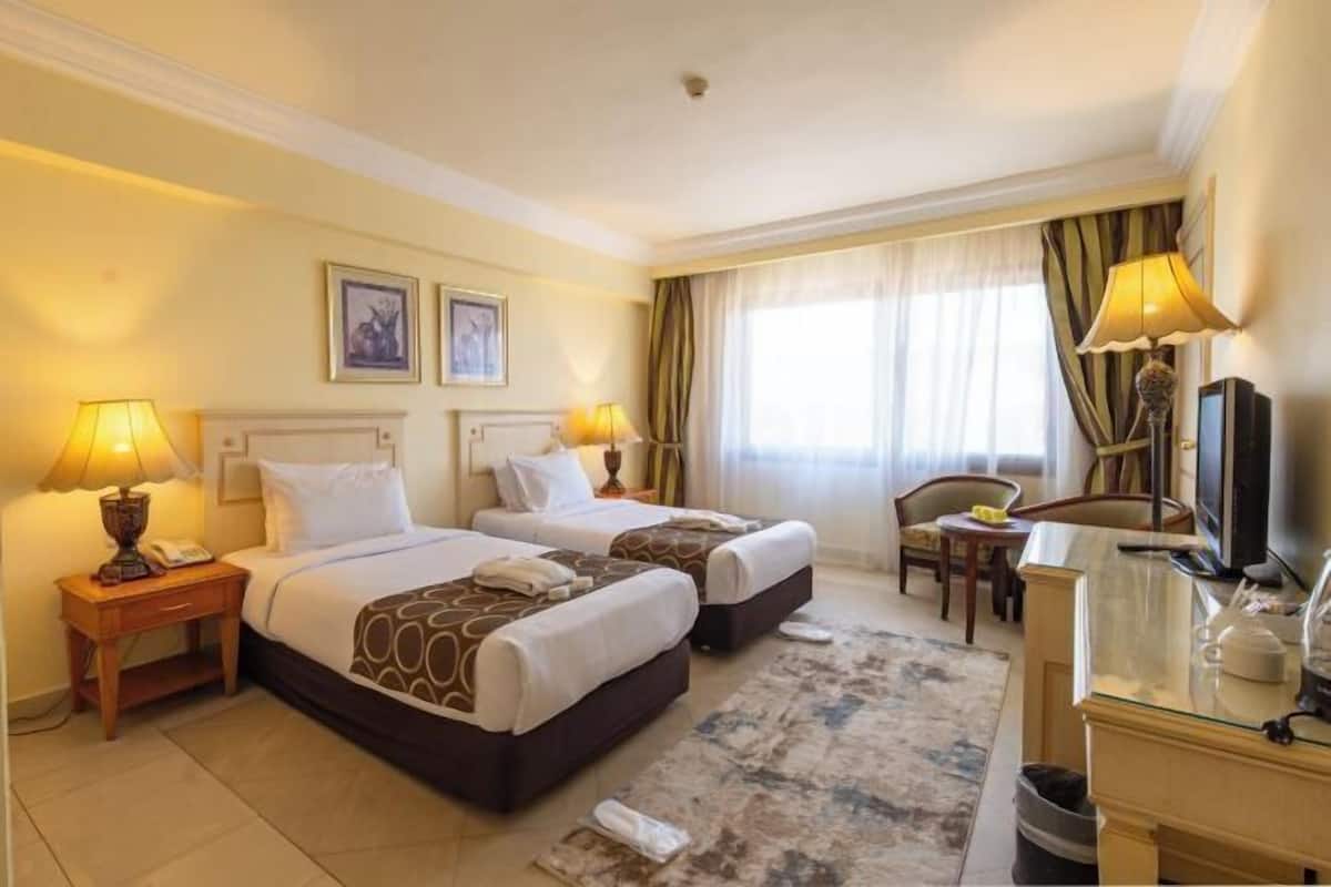 Une chambre d’hôtel avec deux lits, des tables de chevet avec des lampes, un bureau avec une chaise, une télévision et une fenêtre avec des rideaux.