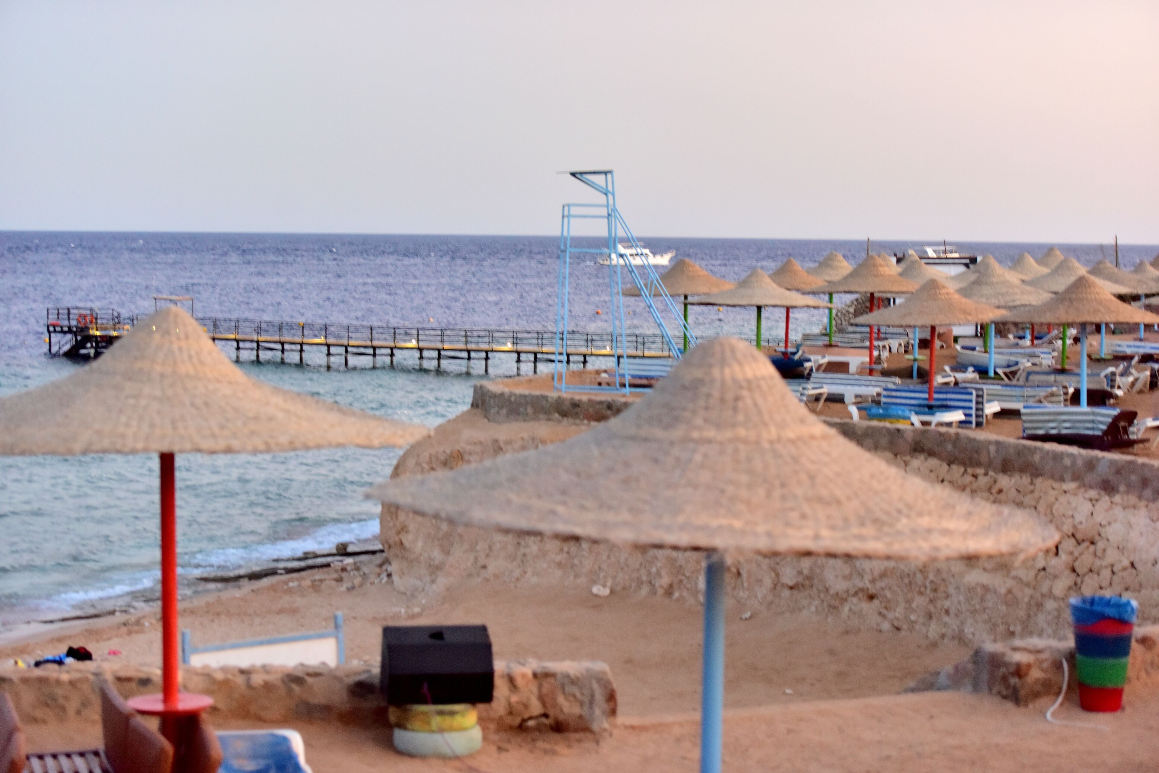 Una playa con sombrillas de paja, un muelle y sillas para tomar el sol.