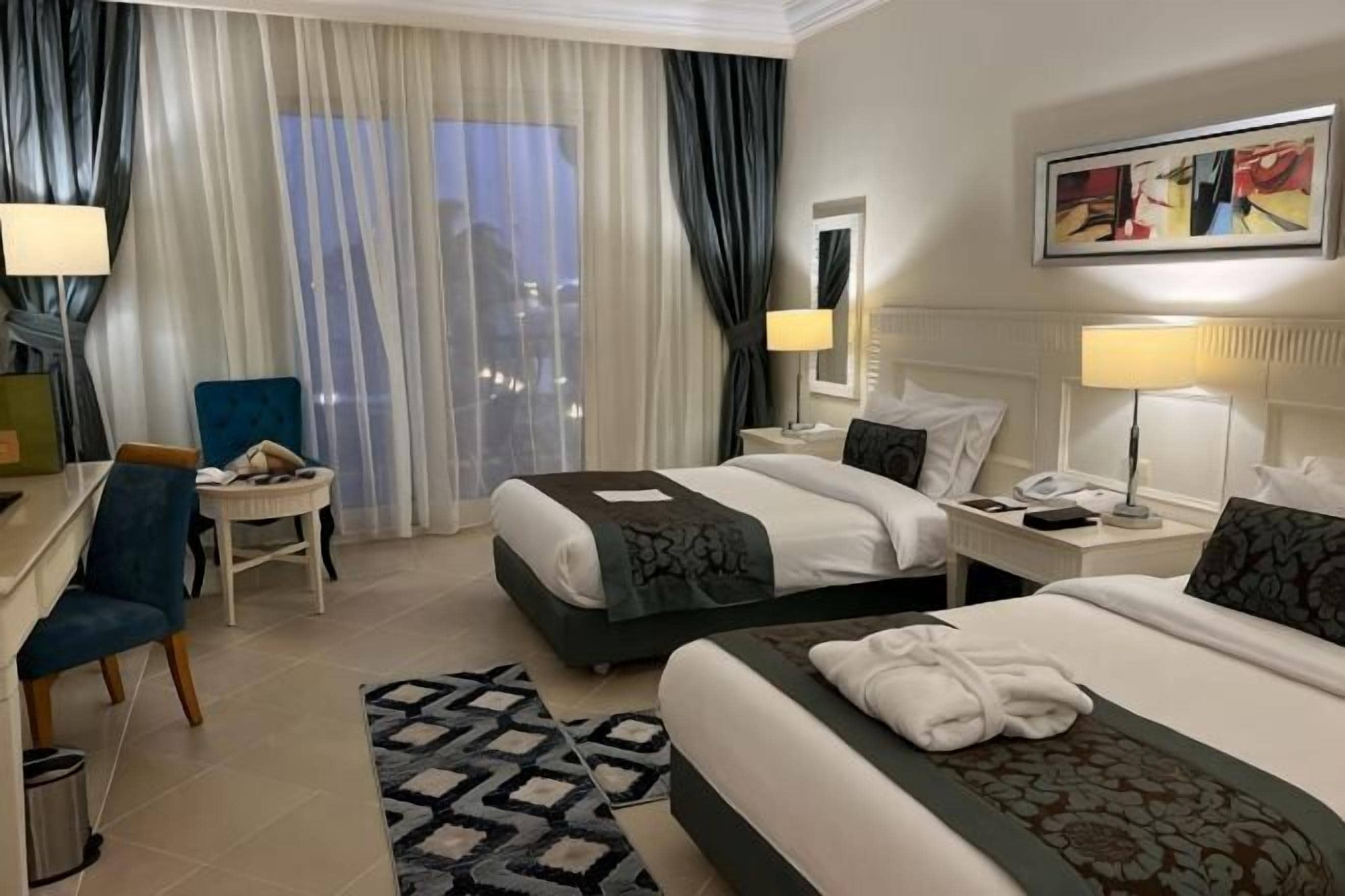 A hotel room with two beds, a desk, a chair, a small table, a lamp, a painting, and a view of the city.