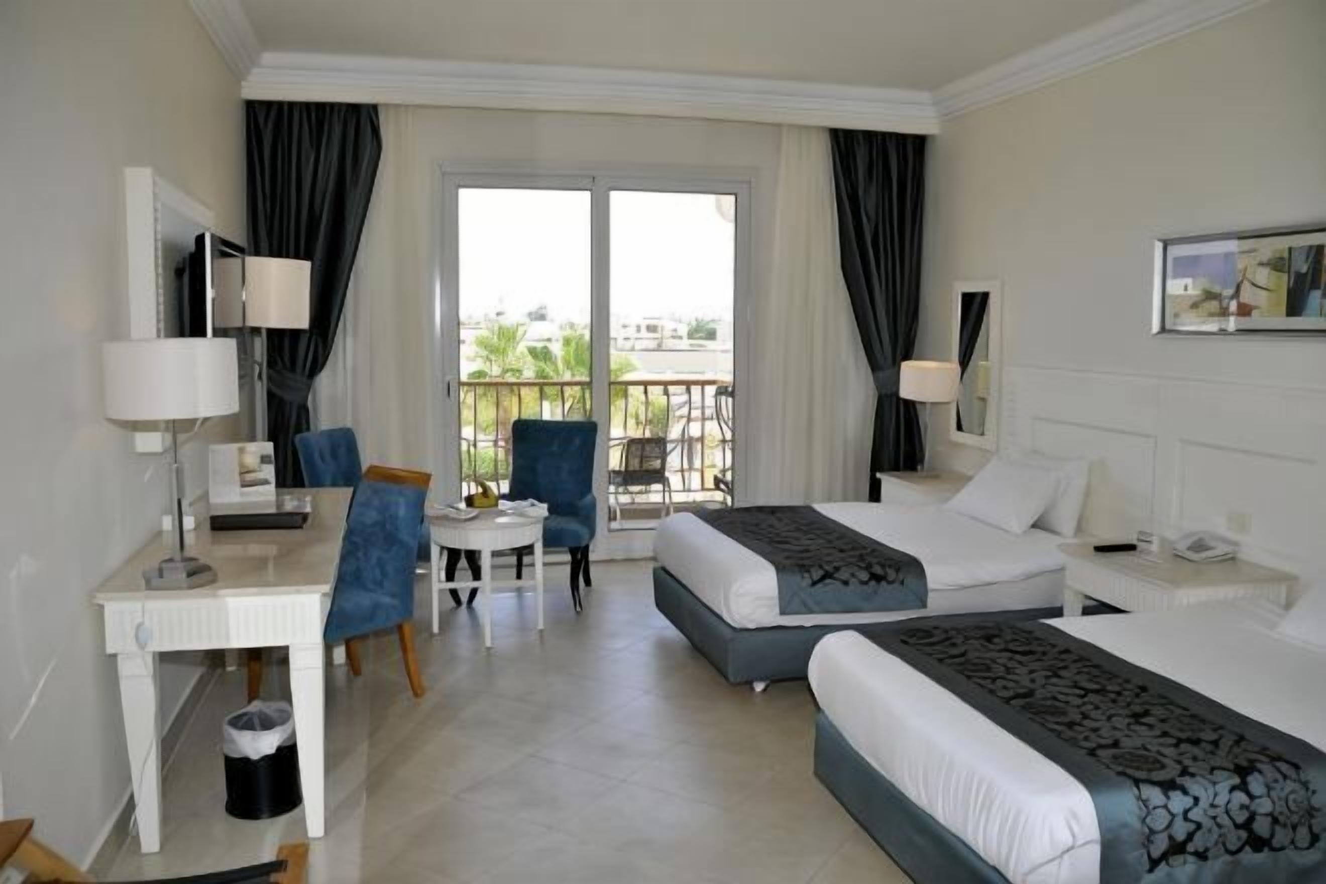 A hotel room with two beds, a desk, a chair, a small table, and a balcony with curtains.