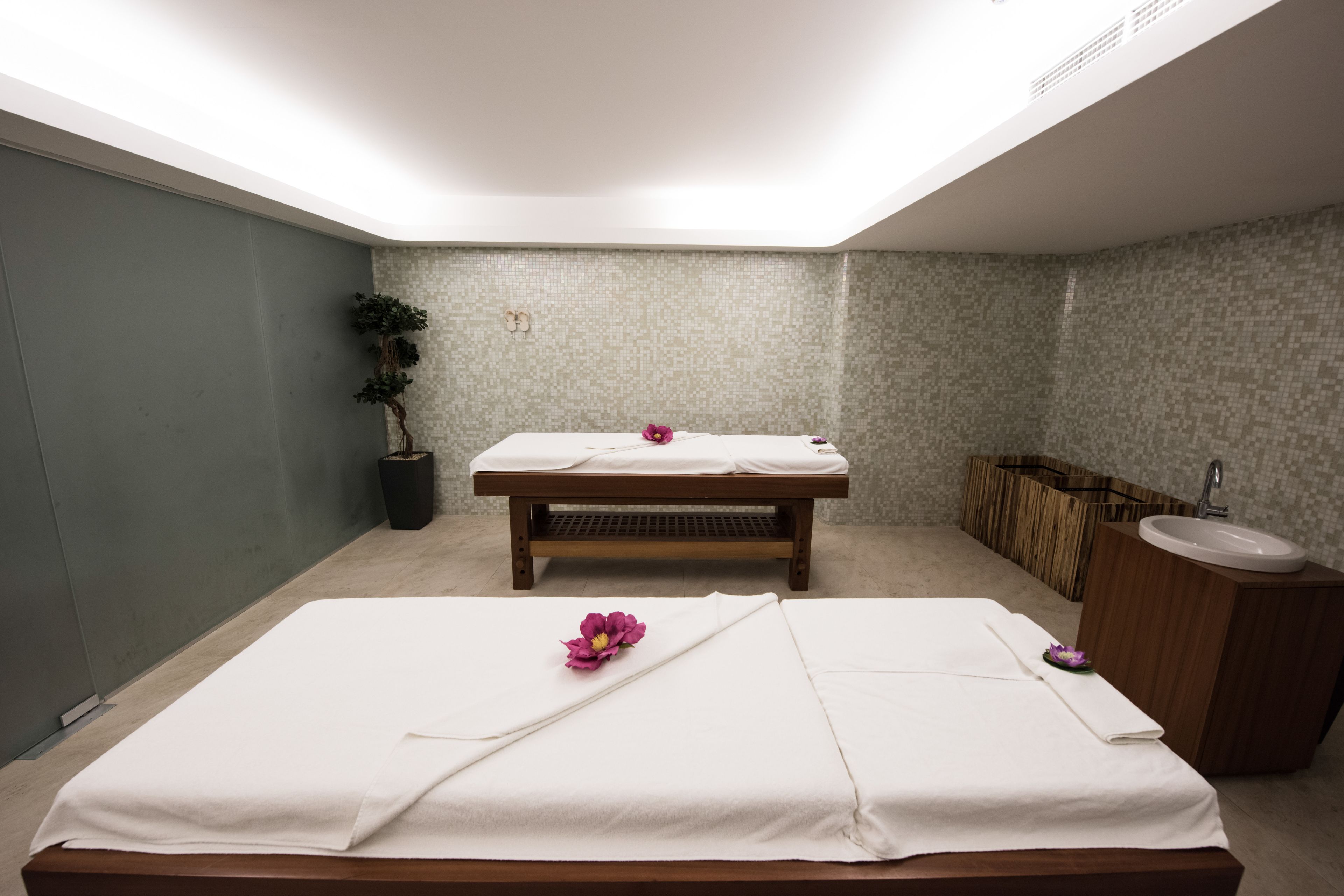 A spa room with two massage tables, a wooden bench, a sink, and a potted plant.
