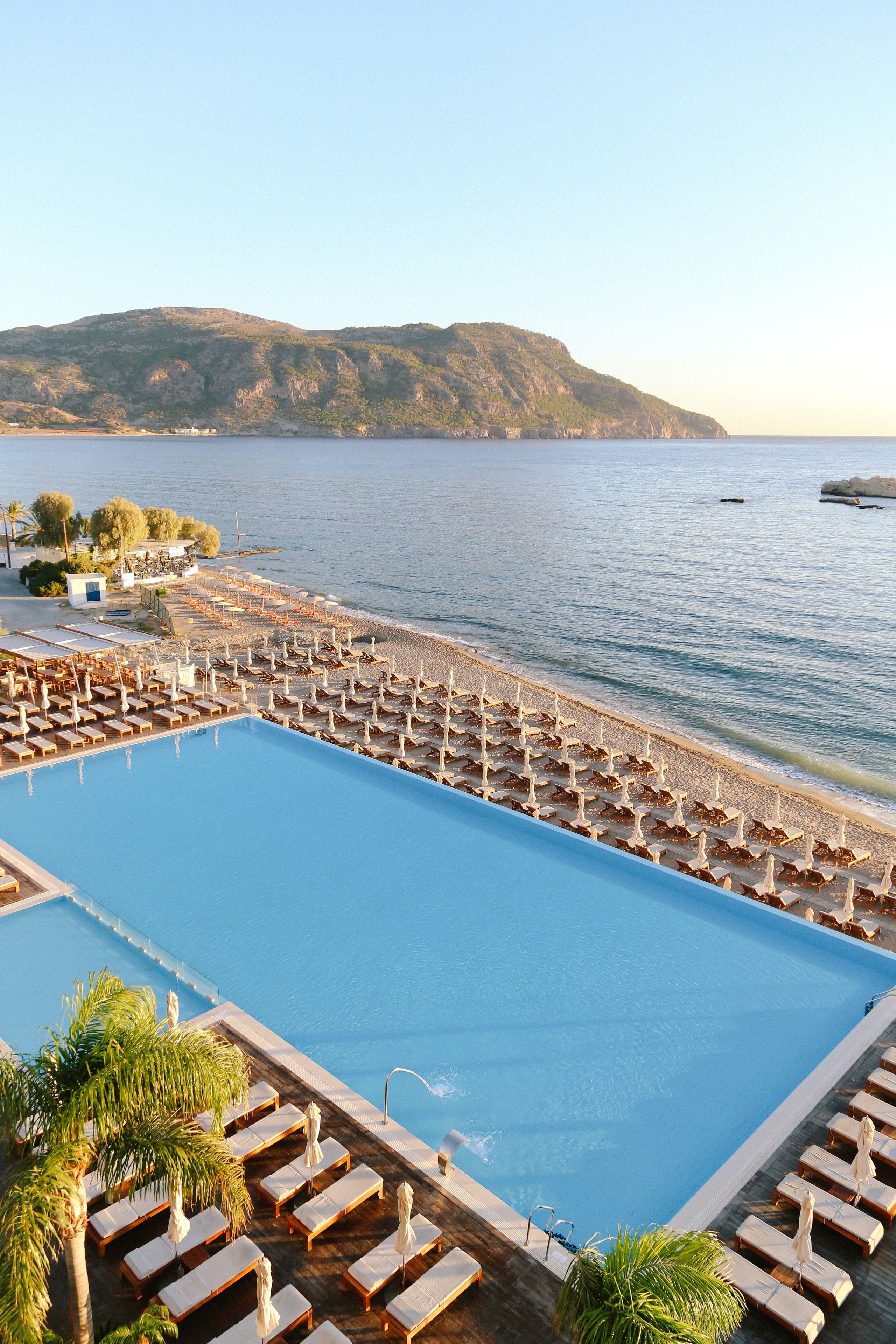 Una piscina con tumbonas y sombrillas, bordeada por una playa y una montaña al fondo.