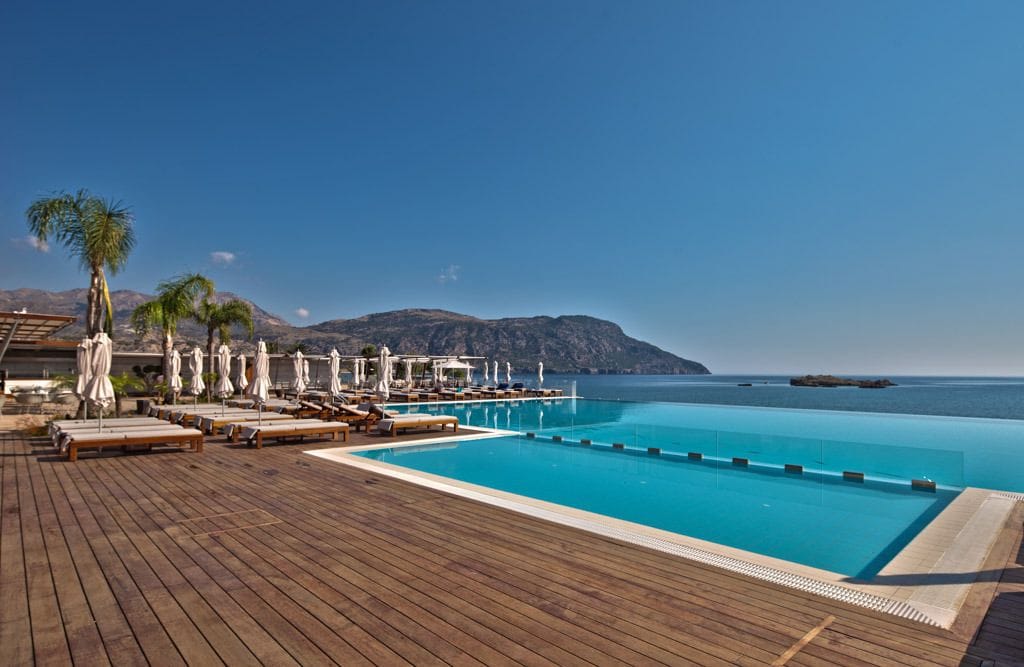 A wooden deck with lounge chairs and umbrellas overlooking a clear blue pool and the sea.