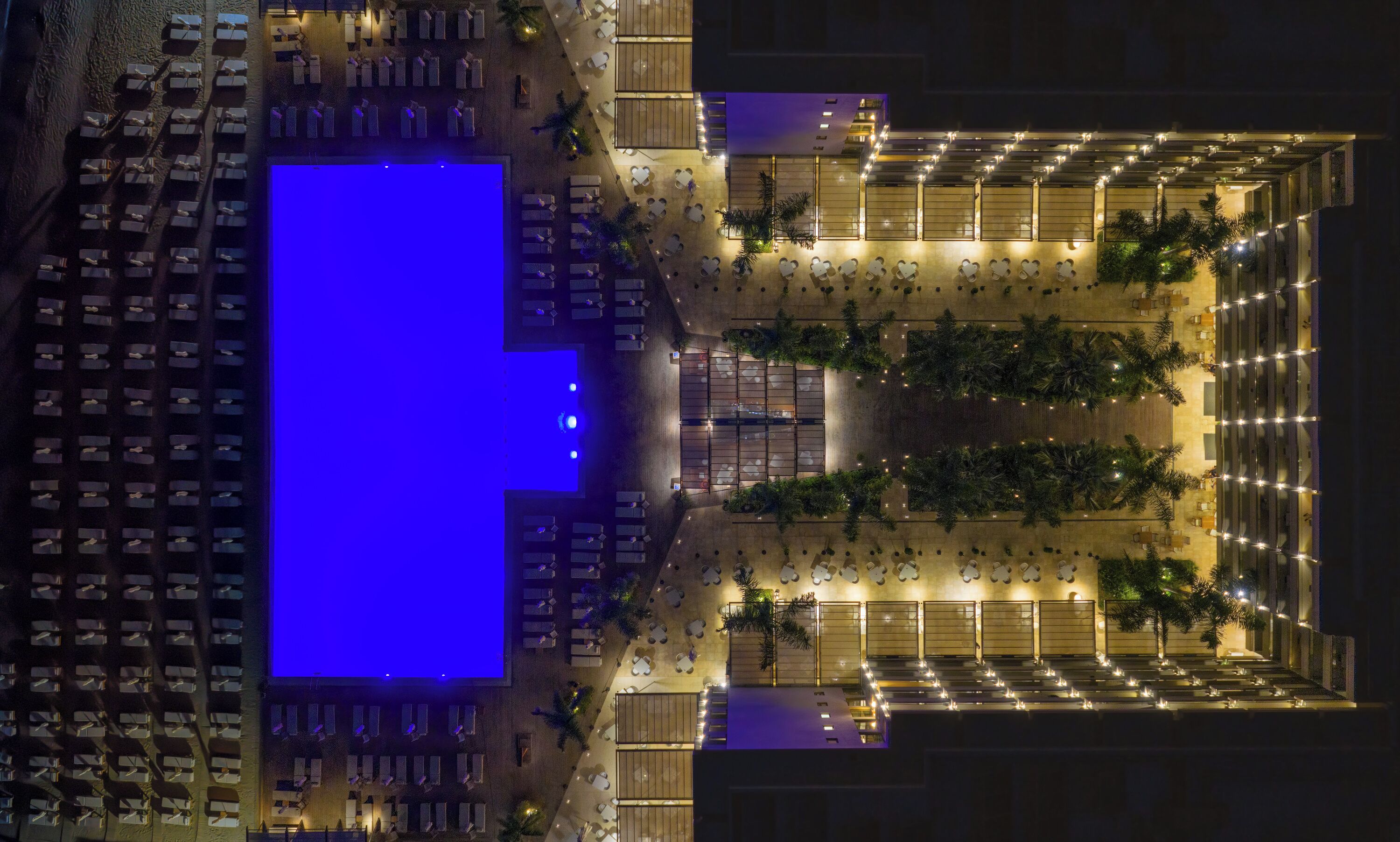 Vista aérea de un hotel con una amplia piscina iluminada en azul, varias zonas de comedor y un área de estar exterior bien iluminada.