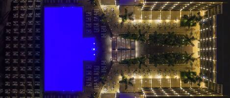 Vista aérea de un hotel con una amplia piscina iluminada en azul, varias zonas de comedor y un área de estar exterior bien iluminada.