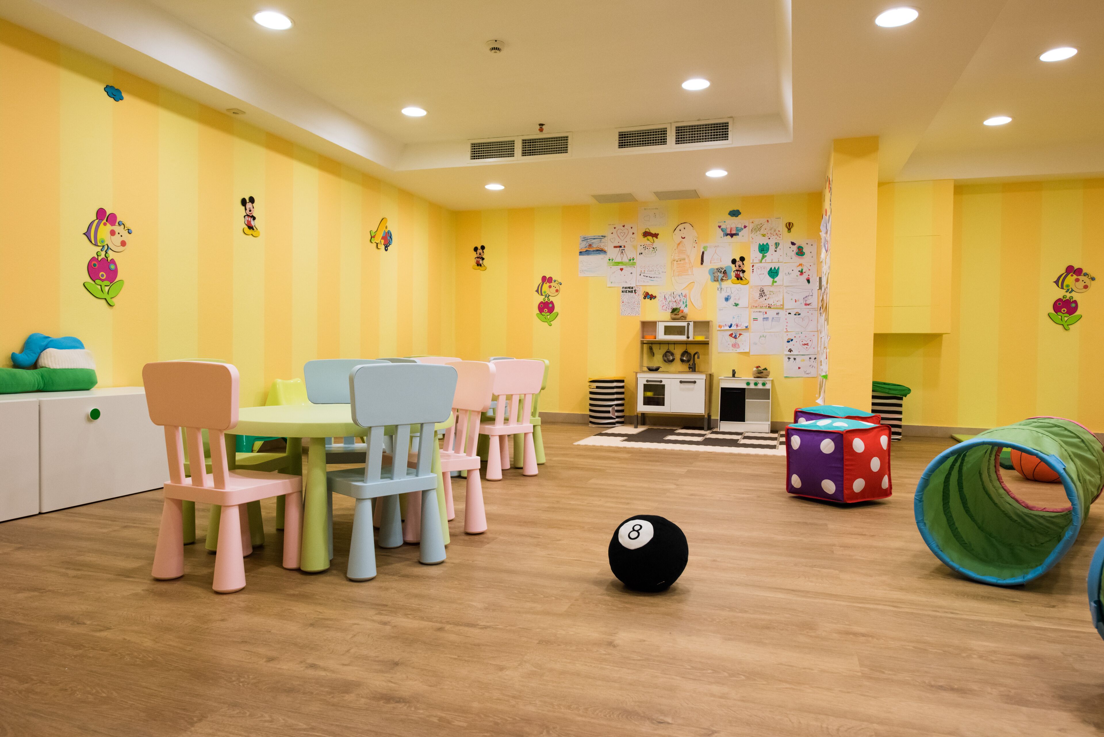 Een helder verlichte kamer met gele muren, meerdere kinderstoelen, een tafel en een speelruimte met een bal en een tunnel.