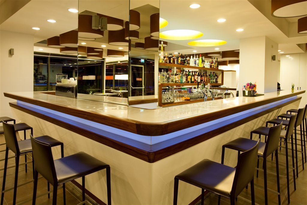 A modern bar area with a long counter, multiple bar stools, and a well-stocked shelf with various bottles.