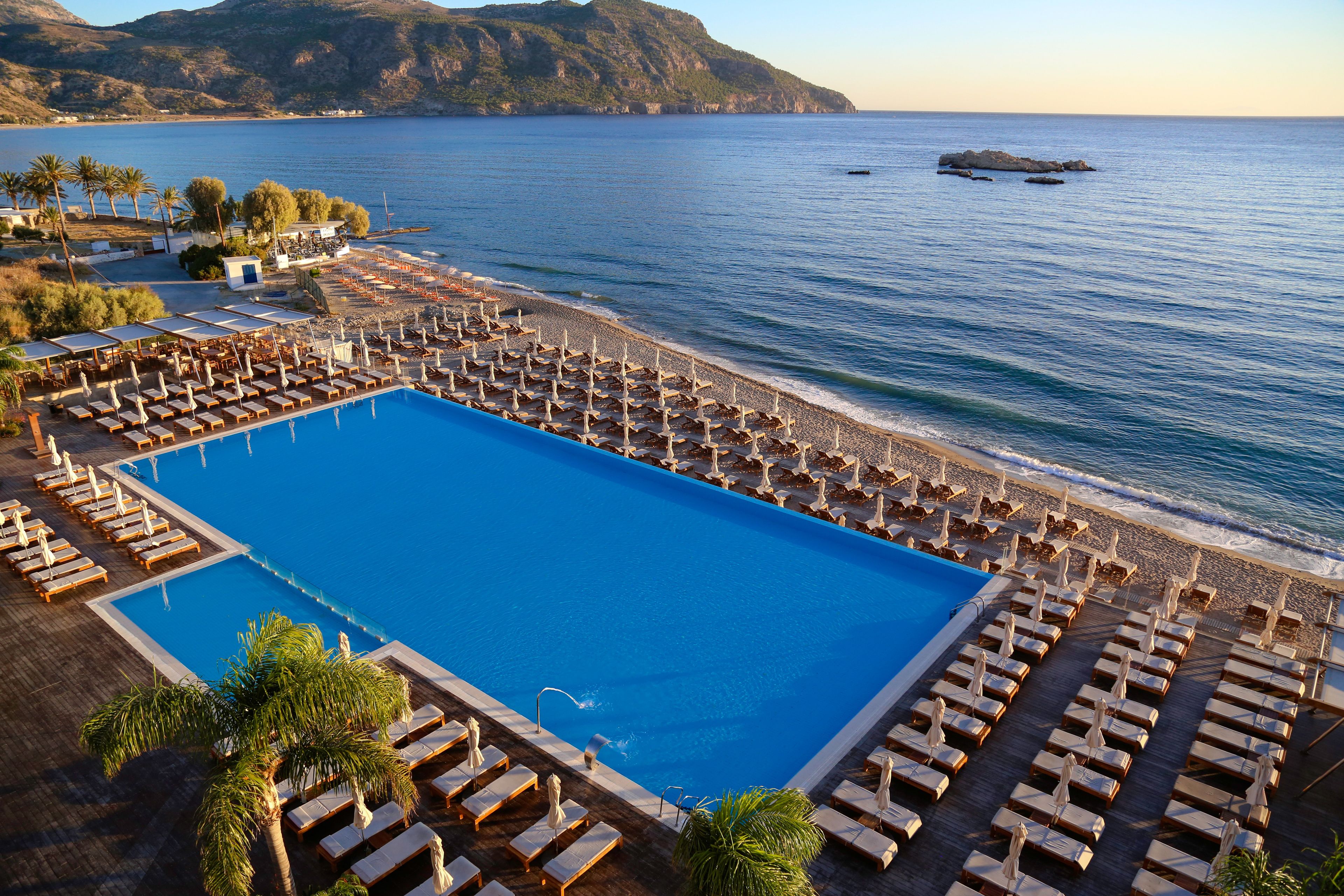 A large rectangular swimming pool adjacent to a beach with rows of sun loungers and umbrellas.