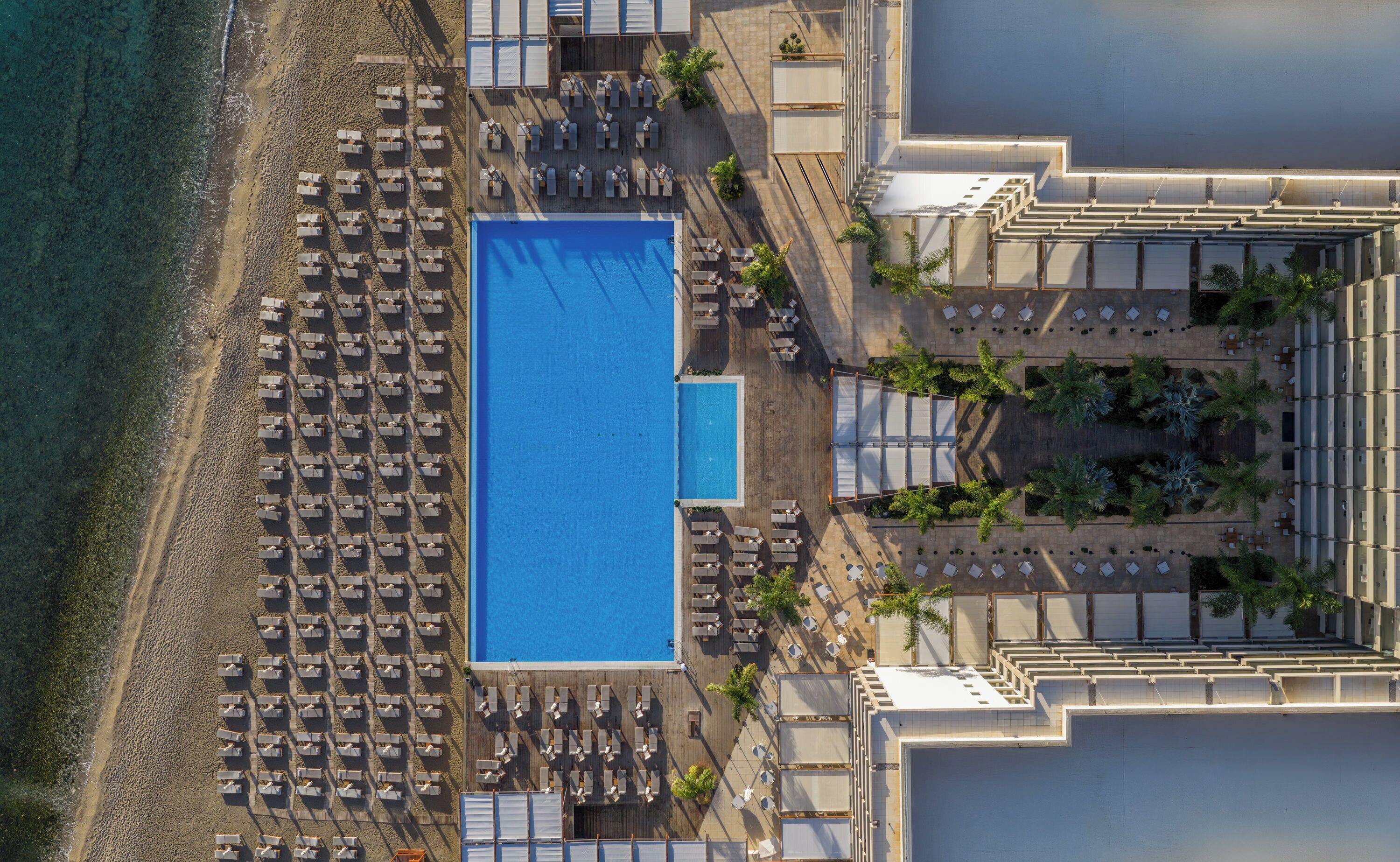 Eine Vogelperspektive eines Resorts mit einem großen rechteckigen Pool, zahlreichen Liegestühlen und Palmen.