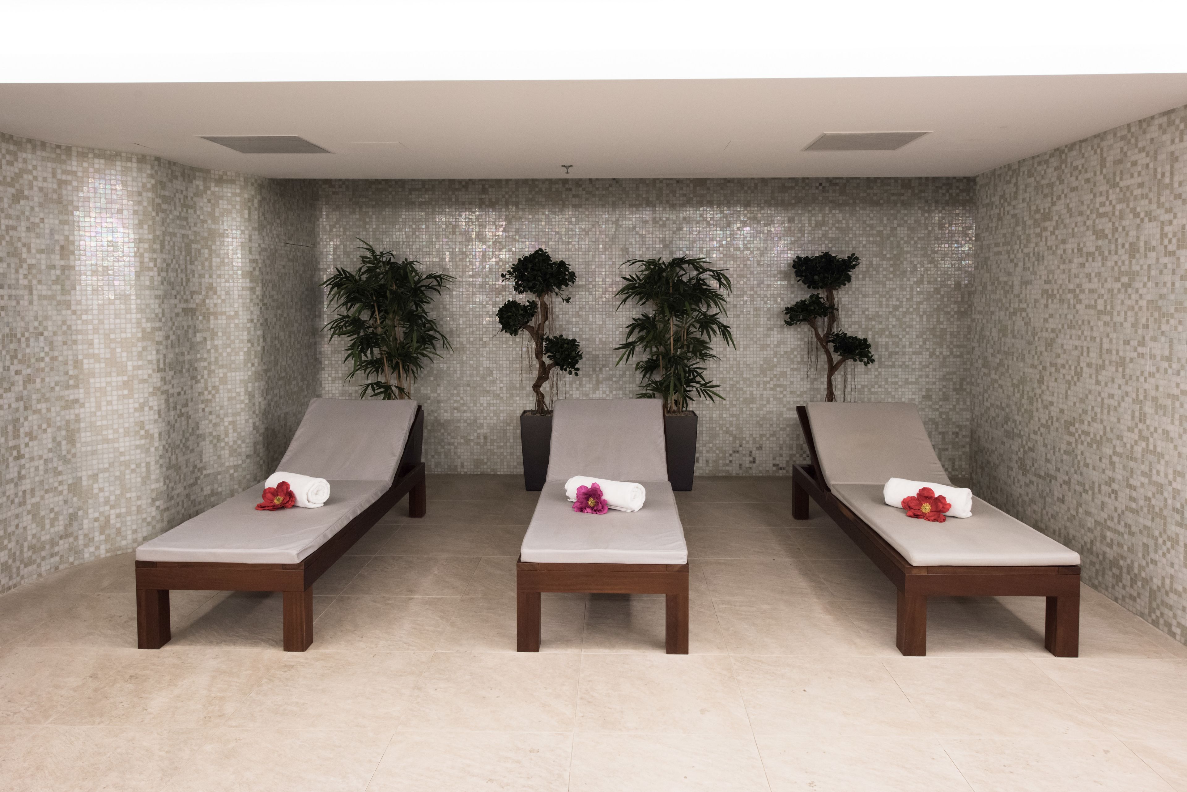 Tres sillones de madera con toallas y flores, frente a una pared de azulejos con plantas en macetas.