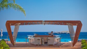 Estructura de pérgola de madera con zona de bar y asientos, con vistas a un mar azul cristalino y una costa rocosa.